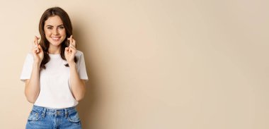 Hopeful woman praying, cross fingers for good luck, making wish, standing over beige background.