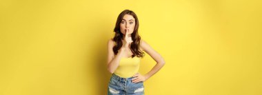 Young woman keeps secret, shows silence quiet, shush gesture, hushing, press finger to lips, standing over yellow background. Copy space