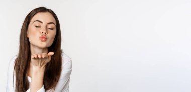 Feminine woman sending air kiss at camera, coquettish flirty pose, kissing, standing over white background. Copy space