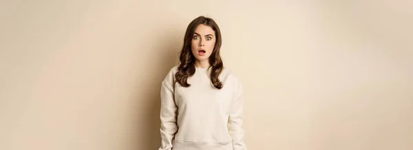 Portrait of shocked brunette woman drop jaw, gasping and staring speechless at camera, beige background. Copy space