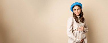 Concept of pregnancy and surrogacy. Young gentle and tender woman with big belly. Smiling pregnant woman posing in trench coat against beige background.