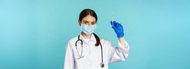 Covid ve sağlık hizmetleri konseptinden aşı. Genç kadın doktor, maske ve eldivenli hemşire aşı yapmak için şırınga kullanıyor, torkuaz arka planda duruyor..