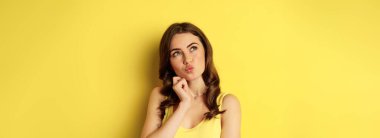 Close up portrait of stylish young beautiful woman thinking, yearning smth, looking up and making choice, standing over yellow background.