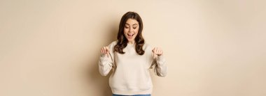 Excited adult woman pointing fingers down and smiling, looking at banner below, showing advertisement, found smth, standing over beige background.