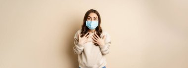 Covid-19, pandemic and quarantine concept. Surprised brunette woman in face medical mask, looking amazed at camera, watching big news, beige background.