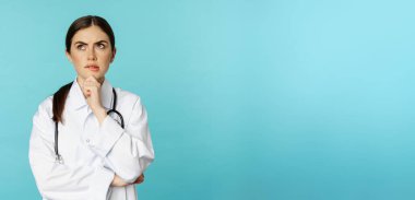 Young female doctor, hospital worker in white coat, thinking and looking away thoughtful, searching solution, standing over toquoise background.