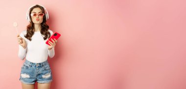 Stylish brunette woman dancing in headphones with mobile phone, listening music in earphones, licking lolipop, standing over pink background.