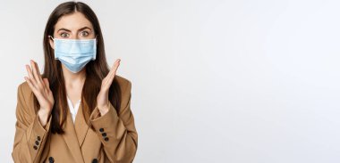 Workaplce ve pandemik konsept. Şok olmuş tıbbi maskeli iş kadını, soluk soluğa, kameraya korkmuş ve endişeli bakıyor, beyaz arka planda duruyor..