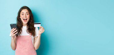 Şaşırmış ve mutlu genç bir kadın plastik kredi kartı ve akıllı telefon gösteriyor, kameraya hayran bakıyor, bahar indirimleri ile online alışveriş yapıyor, mavi arka plan.
