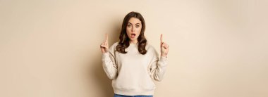 Surprised beautiful girl showing advertisement, pointing fingers on top, store sale or website link up, standing over beige background.