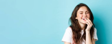 Çekici, kıvırcık saçlı, makyajlı, gülüşlü, gülümsemesini eliyle örten ve mavi arka planda duran genç bir kadını yakından çek..