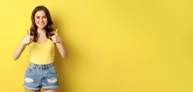 Portrait of stylish modern girl, feminine woman showing thumbs up, recommending gesture, like or approve, praise, standing over yellow background.