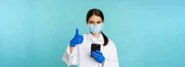 Online doctor and clinic. Young woman in medical face mask, using smartphone for client remote online appointment, showing thumbs up, standing over blue background.