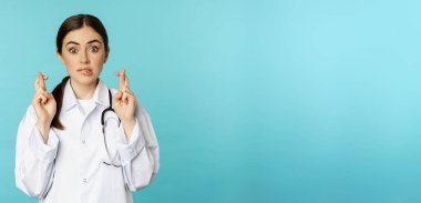 Hopeful woman doctor, lab worker cross fingers and smiling, praying, making wish, anticipating, standing over torquoise background.
