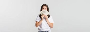 Heyecanlı hayalperest kız elinde para tutuyor ve sol üst köşeye bakıyor, beyaz arka planı düşünüyor..