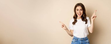 Happy authentic girl smiling, pointing fingers sideways, showing left and right banner, demonstrating promo, standing against beige background.