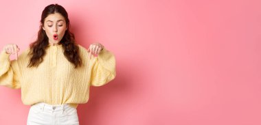 Alttaki tanıtım teklifini kontrol etmek için heyecanlı şirin bir kız, parmakları aşağıda ve diyor ki vay be, reklamlardan etkilenmiş, pembe arka planda duruyor..