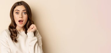 Close up shot of excited woman looking amazed with opened mouth, standing over beige background. Copy space