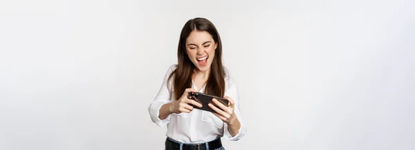 幸せなブルネットの女性は モバイルビデオゲーム 笑顔と見て興奮しました 白い背景の上に立って — ストック写真