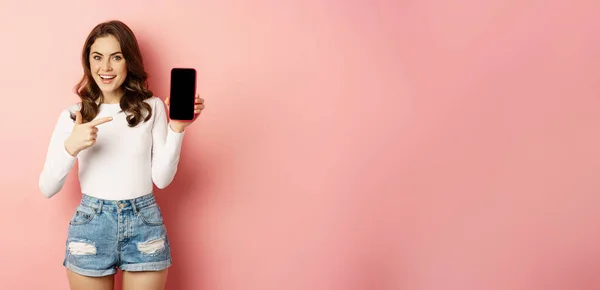 Stylish sexy woman showing mobile phone screen, smartphone app interface and smiling, demonstarting online shop, standing over pink background.