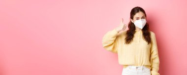 Covid-19, tecrit ve salgın konsepti. Neşeli esmer kız Coronavirus 'u önlemek için medikal solunum cihazı takıyor, başparmağını sallıyor ve evet diyor, pembe arka plana karşı duruyor..