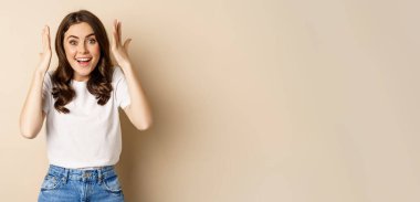Enthusiastic adn surprised woman winning, dancing and celebrating, standing in tshirt over beige background.