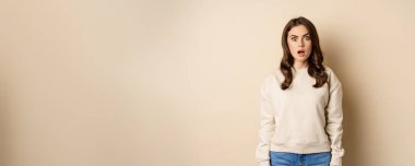Portrait of shocked brunette woman drop jaw, gasping and staring speechless at camera, beige background. Copy space