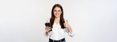 Mutlu gülümseyen kurumsal kadın, baş parmak gösteren kadın model, akıllı telefon tutan, cep telefonu kullanan, beyaz arka planda duran.