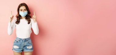 Portrait of young stylish woman in casual summer clothes, wearing face medical mask, pointing fingers up, showing promo offer, standing against pink background.