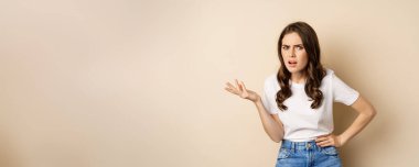 Confused woman looking clueless and annoyed, cant understand, asking whats problem, standing over beige background.