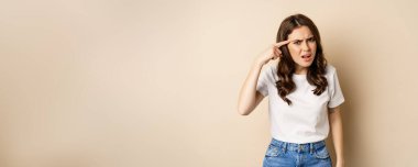 Annoyed and angry woman pointing finger at head, scolding someone, standing over beige background.