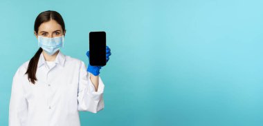 Young woman doctor in medical face mask and hospital uniform, showing mobile phone app, screen interface, online clinic concept, standing over torquoise background.