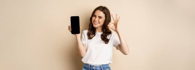 Smiling happy girl showing mobile phone screen, app on smartphone and okay, recommending online store, shopping application, beige background.