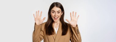 Enthusiastic businesswoman in suit, raising hands palms up, number ten dozen gesture and smiling, standing over white background.