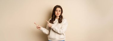 Skeptical girl looking hesitant, pointing fingers right at logo, promo text, standing over beige background.