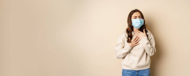 Covid-19 and health concept. Sick woman in medical face mask coughing, feeling ill with sour throat, standing over beige background.