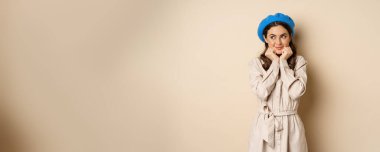 Portrait of beautiful woman in trendy outfit, touching her face and looking dreamy, yearning smth, standing against beige background. People concept
