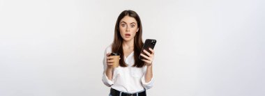 Cep telefonunu kontrol ettikten sonra şaşıran, elinde kahve fincanı tutan beyaz arka planda duran genç bir kadının portresi..