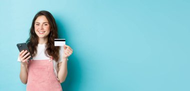Çevrimiçi ödeme yapan genç bir kadın, elinde kredi kartı ve akıllı telefonla online alışveriş yapıyor, kameraya tatmin olmuş görünüyor, mavi arka planda duruyor..