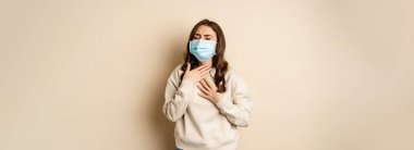 Covid-19 and health concept. Sick woman in medical face mask coughing, feeling ill with sour throat, standing over beige background.