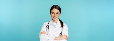 Medical staff and doctors concept. Young smiling female doctor, healthcare worker in white coat and stethoscope, looking confident, waiting patients, blue background.