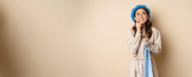 Image of modern beautiful woman posig with shopping gift bag, smiling and looking up thoughtful, thinking, standing over beige background.