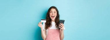 Şaşırmış ve mutlu genç bir kadın plastik kredi kartı ve akıllı telefon gösteriyor, kameraya hayran bakıyor, bahar indirimleri ile online alışveriş yapıyor, mavi arka plan.