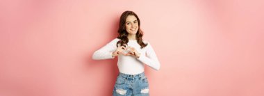 Romantic girl showing heart sign, love like gesture and smiling. Stylish modern woman posing on valentines pink background.