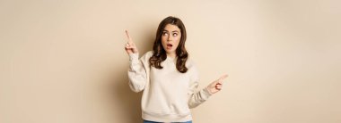 Confused girl shopper pointing sideways, making choice, decision between two variants, beige background. Copy space