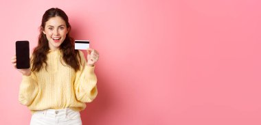 Teknoloji ve online alışveriş. Plastik kredi kartı ile boş akıllı telefon ekranı gösteren genç ve çekici bir kadın model. Banka hesabı ya da başvuru formunu pembe arka planda duruyor..