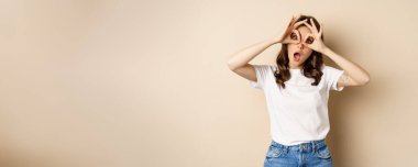 Funny brunette woman having fun, showing finger glasses gesture and fool around, posing over beige background.