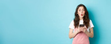 Güzel esmer kızın portresi, elinde cep telefonu, elinde cep telefonu, etkileyici yüzü ve mavi arka planı olan kameraya 