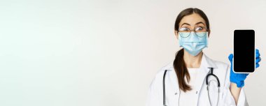 Online medical help concept. Surprised female doctor in face mask, looking amazed while showing mobile phone app, smartphone website, white background.