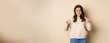 Excited adult woman pointing fingers down and smiling, looking at banner below, showing advertisement, found smth, standing over beige background.